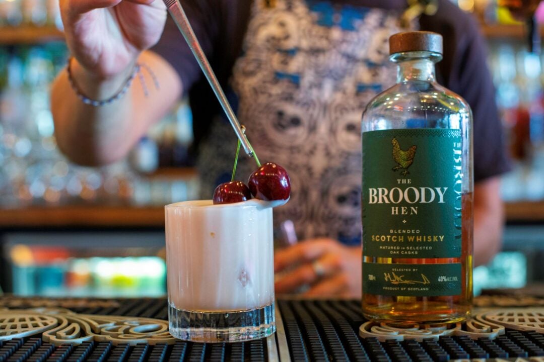 Bartender preparing a drink: on the bar is a milky-looking drink, decorated with 2 cherries, and a bottle of whisky.